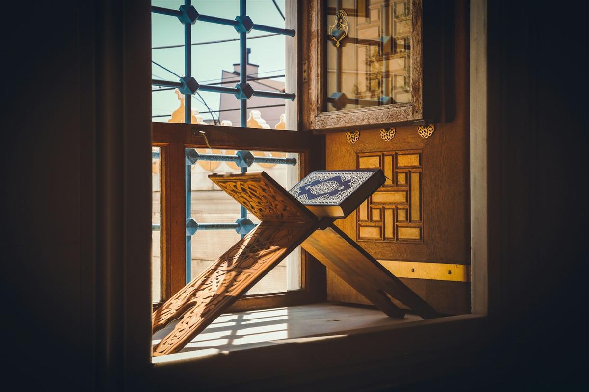 An open Quran resting on a wooden stand in warm sunlight by a window